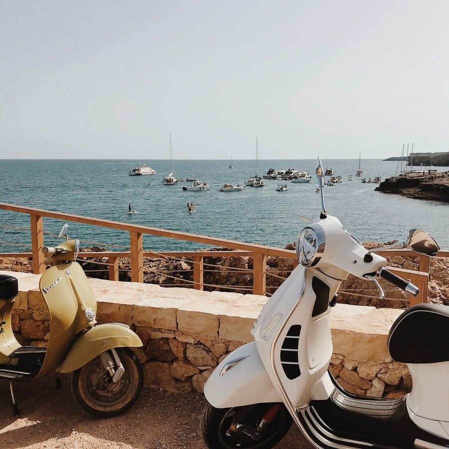 Two Vespas parked on sandy ground beside a low wall above the sea